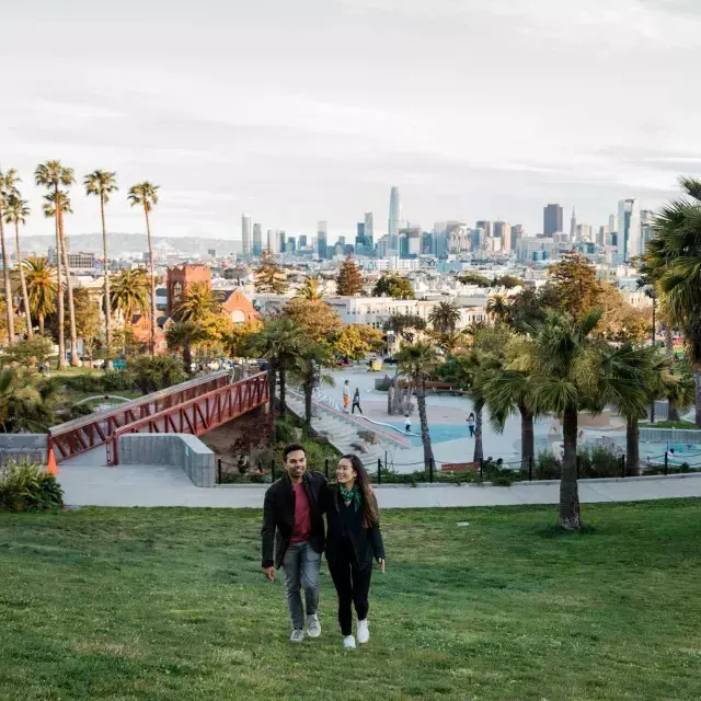 Um casal caminha em direção à câmera com Dolores Park e o horizonte 来自贝博体彩app atrás deles.